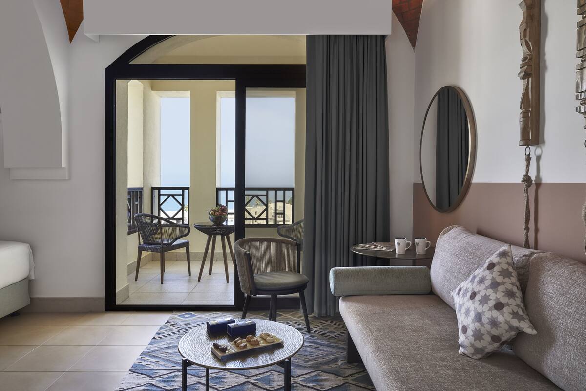 A modern hotel room with a sofa, a small round table with a tray of snacks, a balcony with chairs, and a large mirror on the wall.