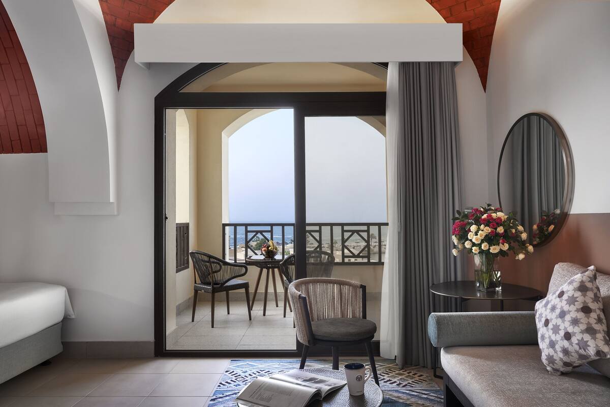 A hotel room with a balcony, a sofa, a chair, a table with flowers, and a mirror.