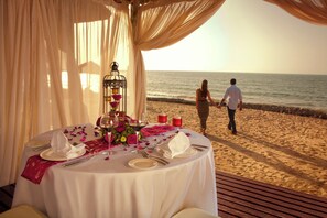 A romantic outdoor dining setup with a table set for two, overlooking a beach. - The Cove Rotana Resort (Ras Al Khaimah)