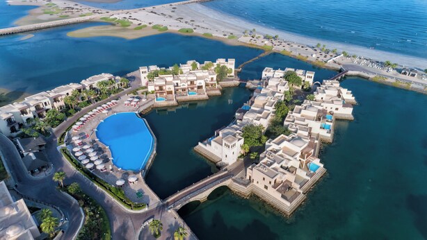 Vista aérea de un complejo turístico costero con varias construcciones, una piscina central y un puerto deportivo.