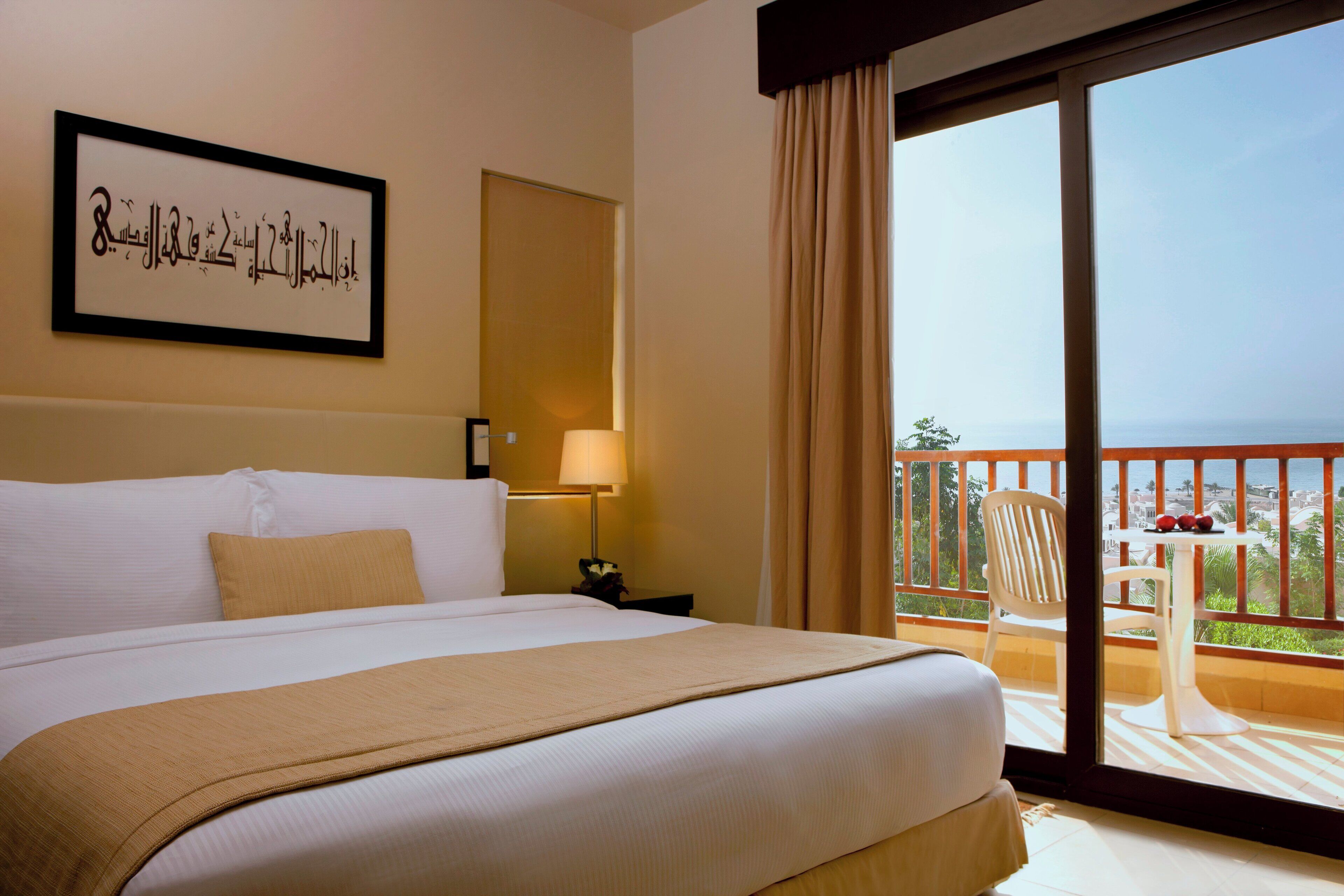 Habitación de hotel con una cama grande, una obra de arte enmarcada en la pared, una mesita de noche con lámpara y un balcón con mesa y sillas.