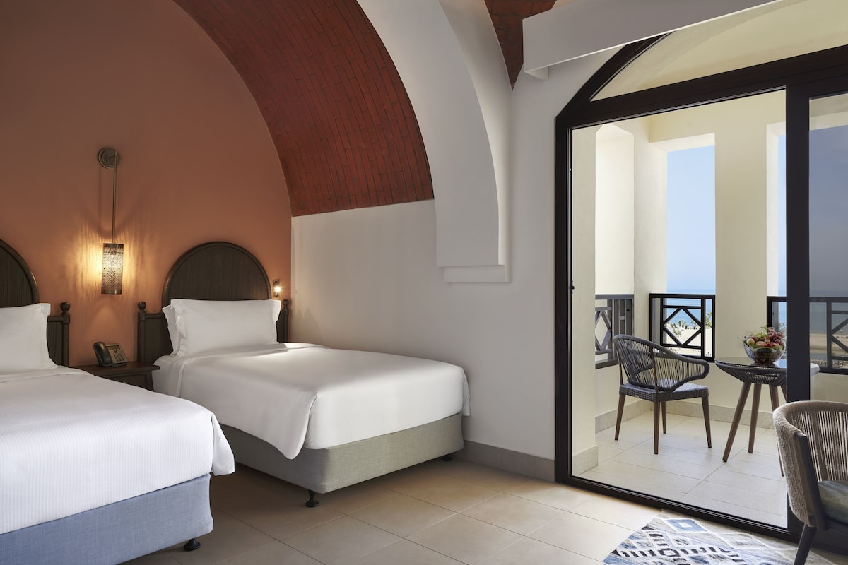 A hotel room with two beds, a balcony with a chair and table, and a view of the outside.