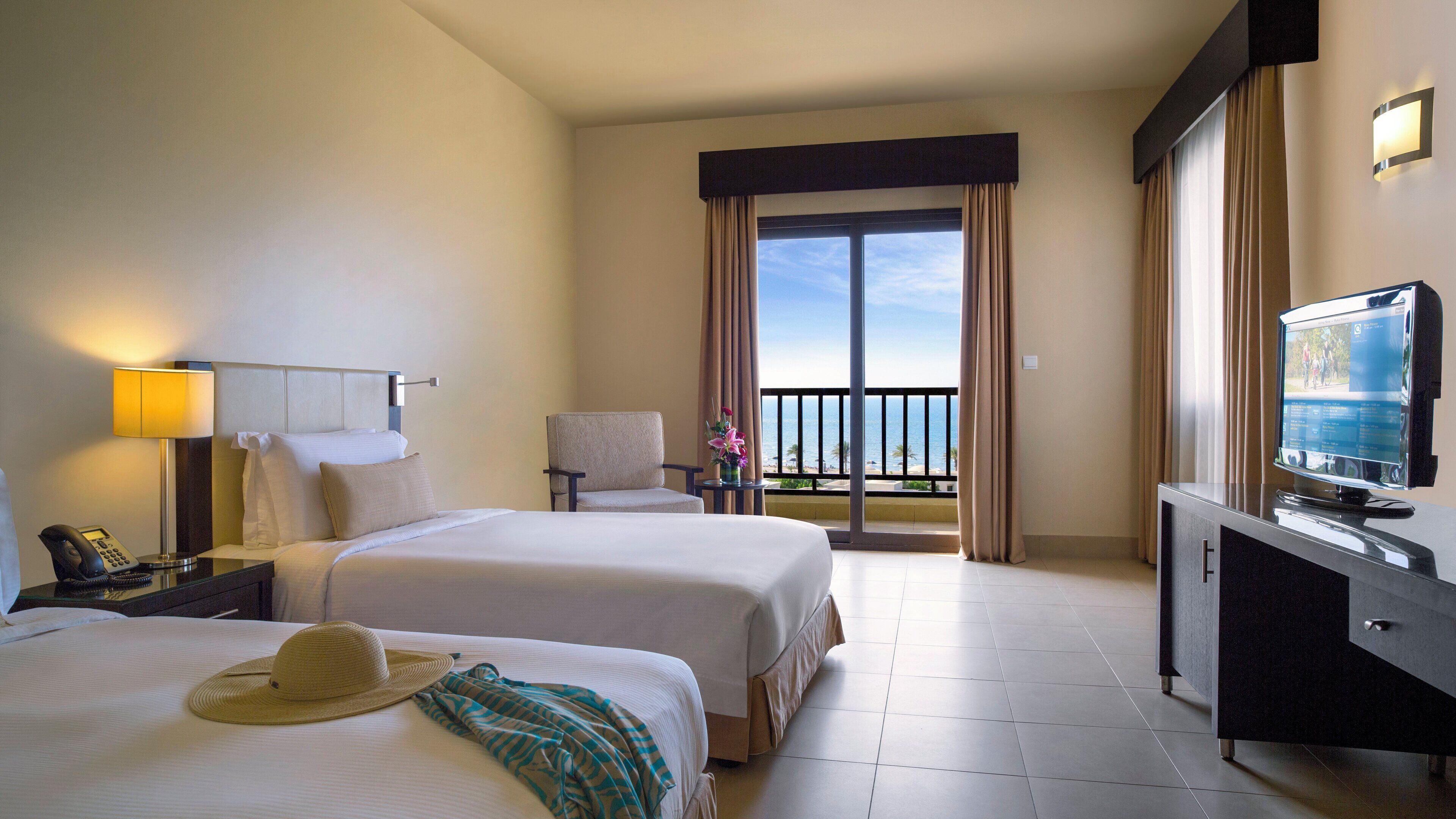 Ein Hotelzimmer mit zwei Betten, einem Schreibtisch mit Computer, Telefon, Fernseher und einem Balkon mit Blick auf den Strand.