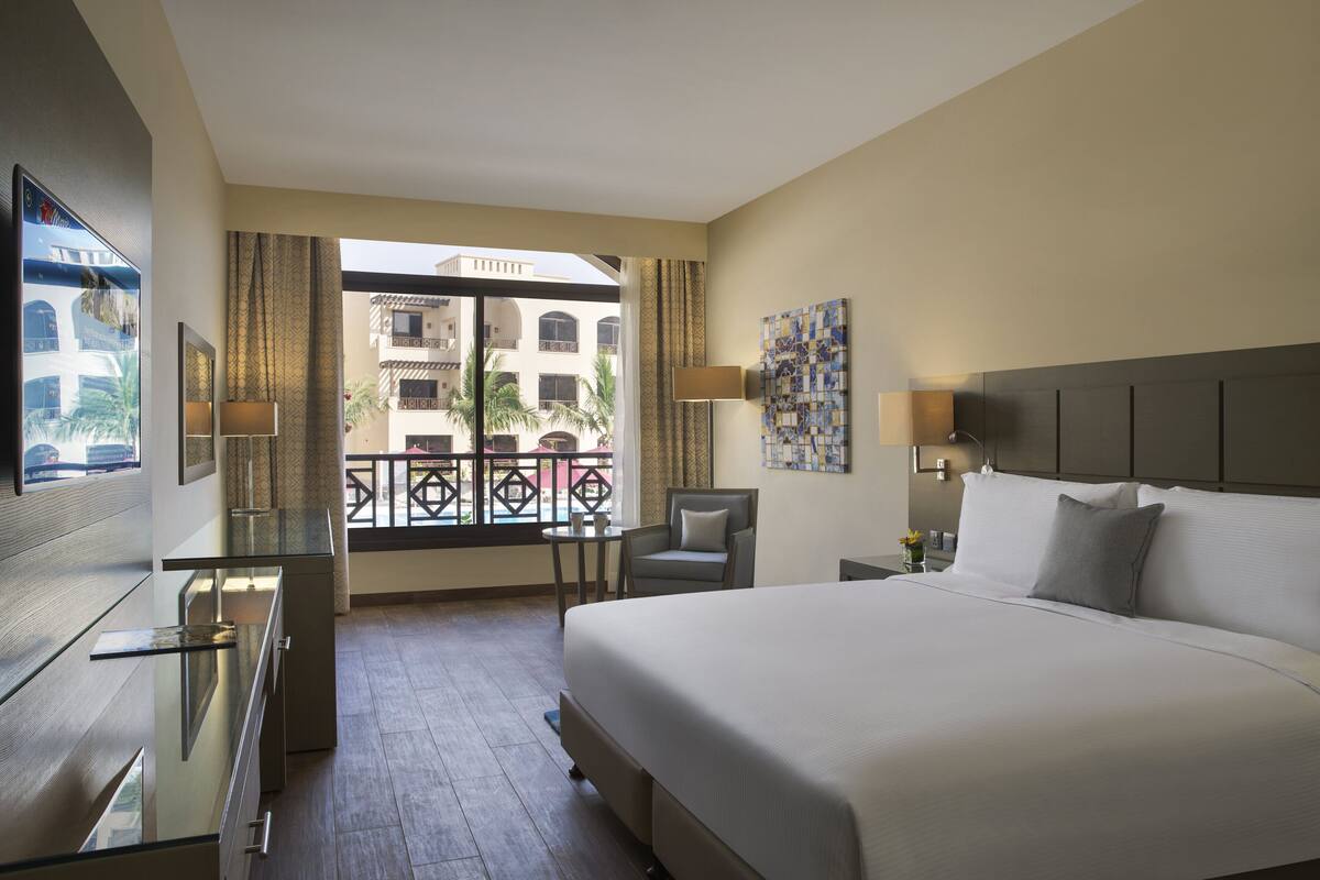 A hotel room with a large bed, a desk, a chair, and a balcony with a view of buildings.