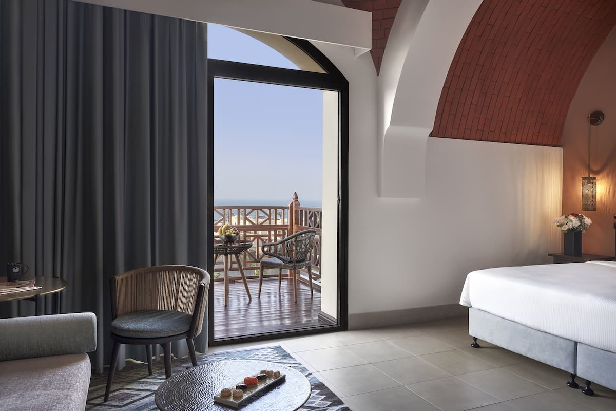 A hotel room with a large bed, a seating area with a chair and table, and a balcony with a view of the sea.