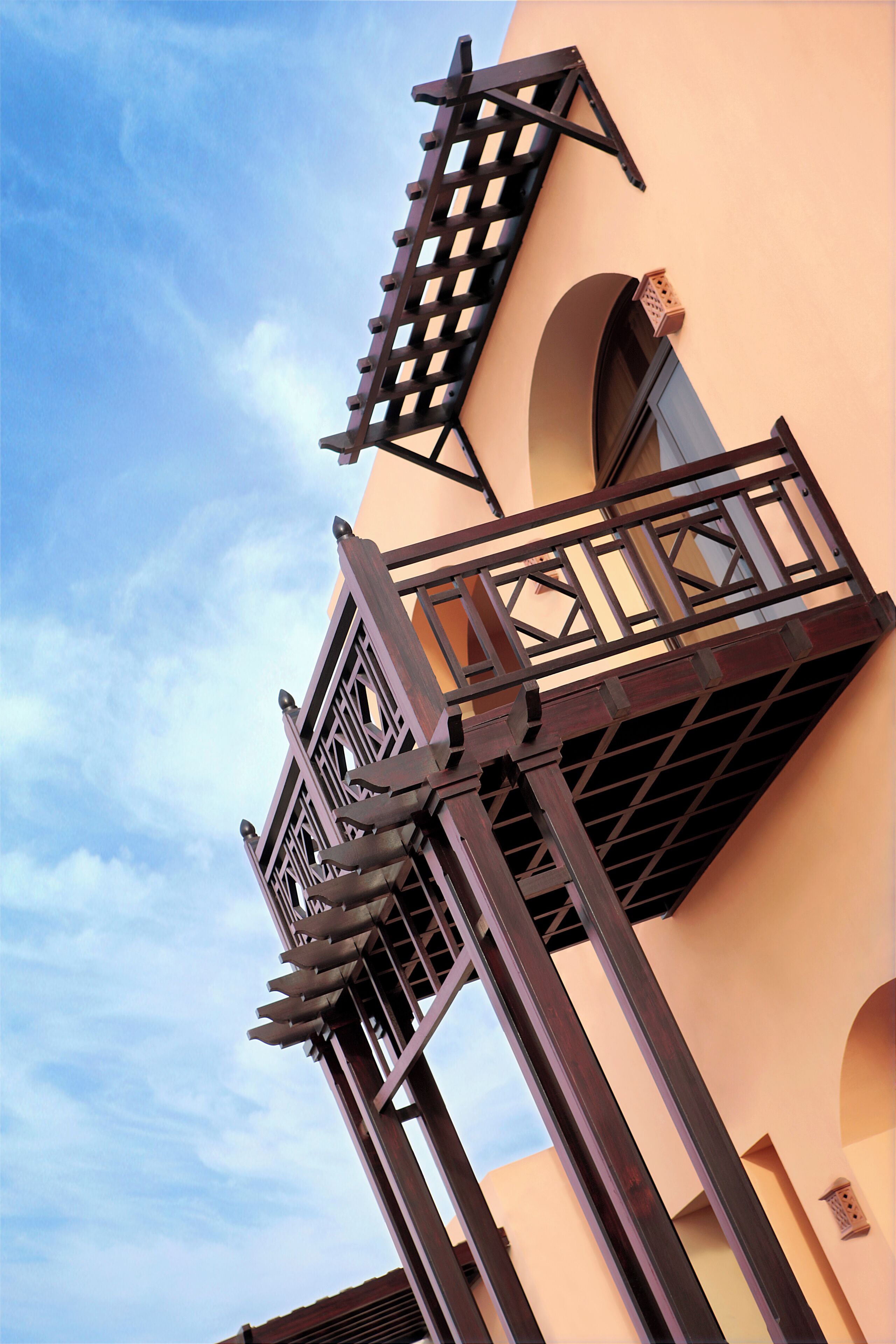 A balcony with a lattice design, attached to a building with arched windows and a peach-colored facade.