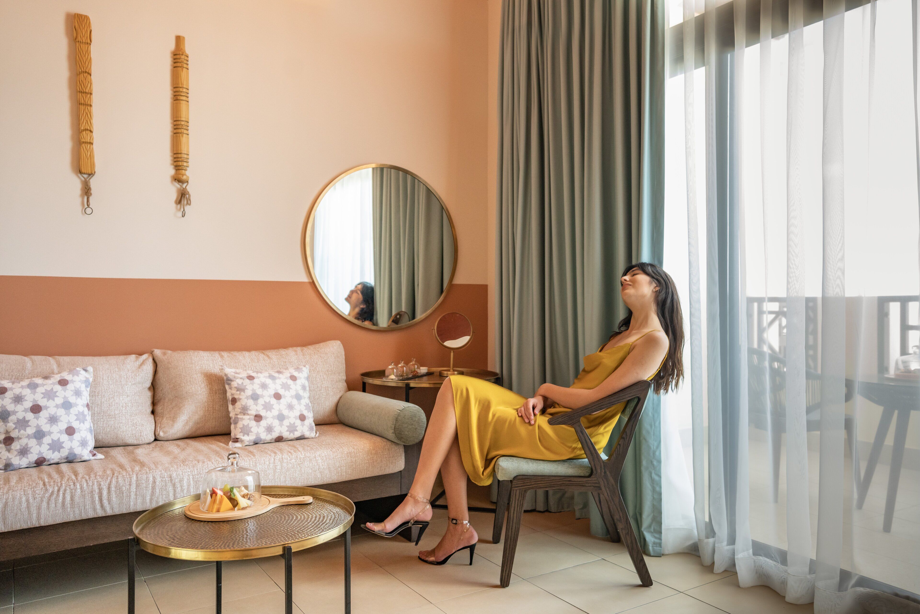 Mulher sentada em uma cadeira em um quarto com um sofá, um espelho redondo e uma mesa com uma bandeja de comida.