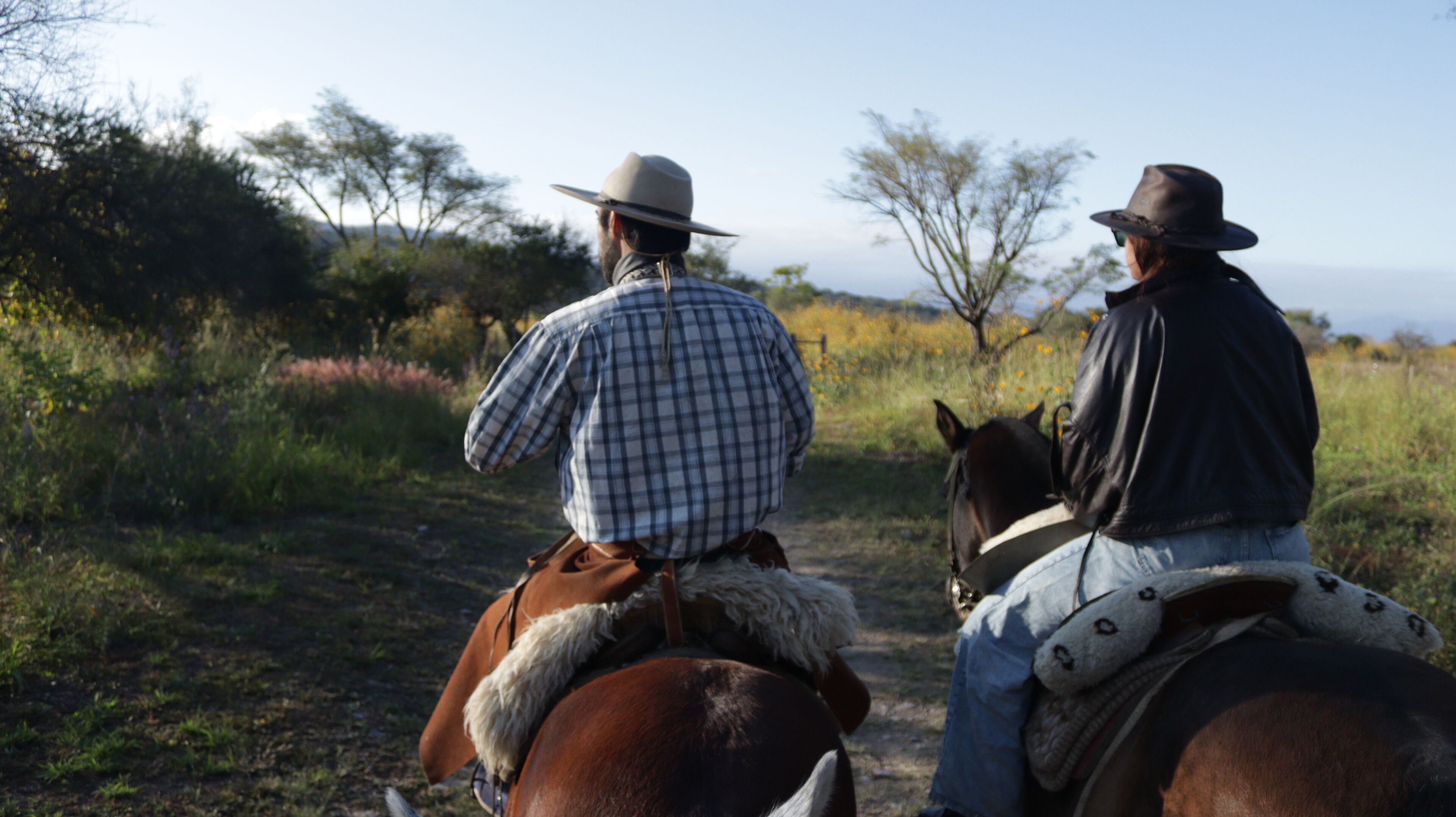 Dos personas montando a caballo por un sendero de tierra rodeado de vegetación.