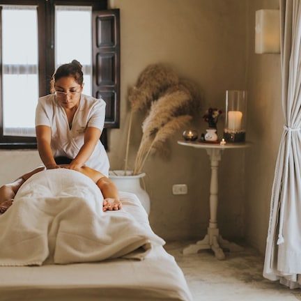 A massage therapy session in a room with a bed, a window with shutters, a small table with candles, and a curtain.