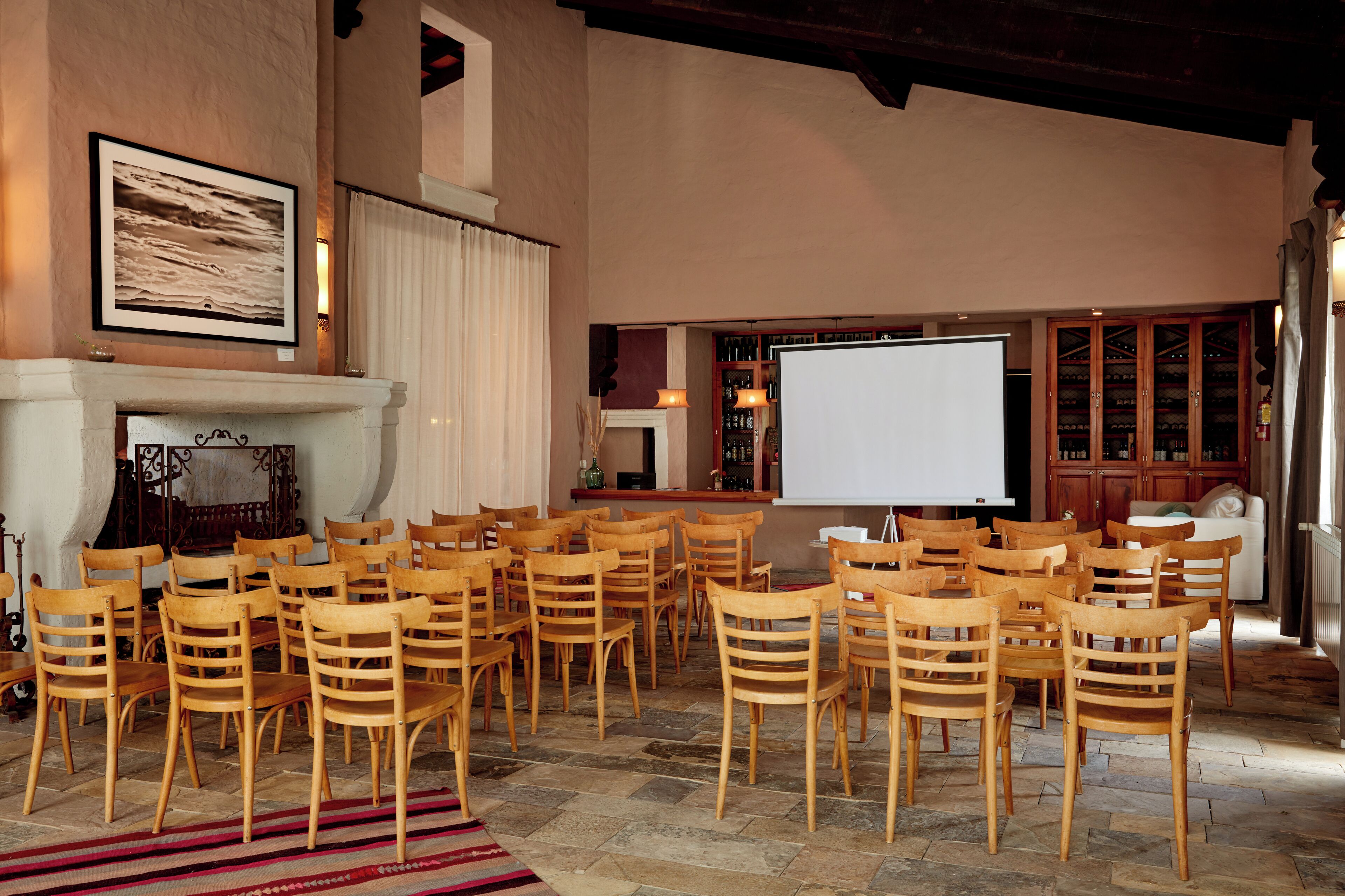 A conference room with wooden chairs arranged in rows, a fireplace, a projector screen, and a wine cabinet.