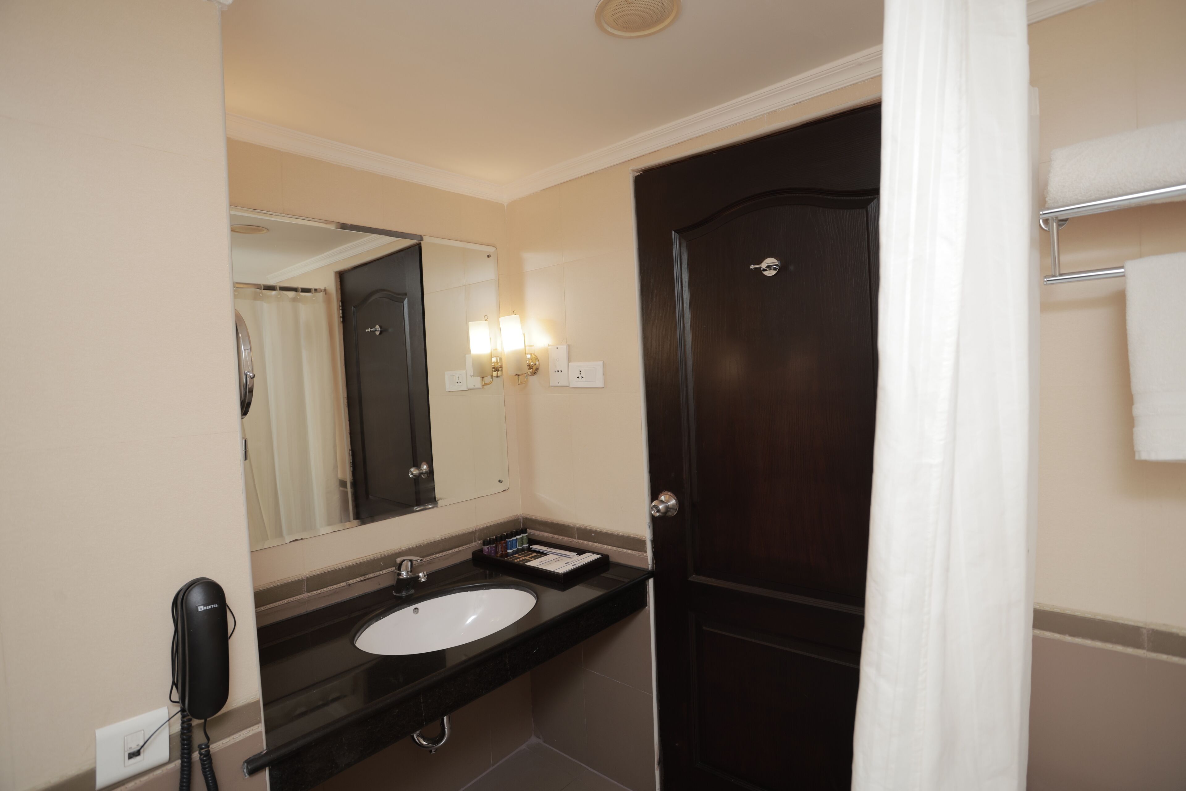 A hotel bathroom with a dark wooden door, a white sink, a mirror, and a telephone.