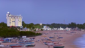 A beachfront hotel with multiple floors, a beach in the foreground with numerous boats, and a cityscape in the background. - The Quilon Beach Hotel and Convention Center (Kollam)