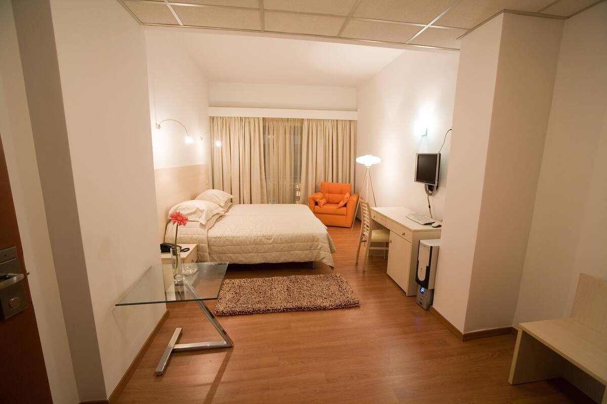 A hotel room with a bed, a desk with a computer, a chair, a TV, and a glass table.