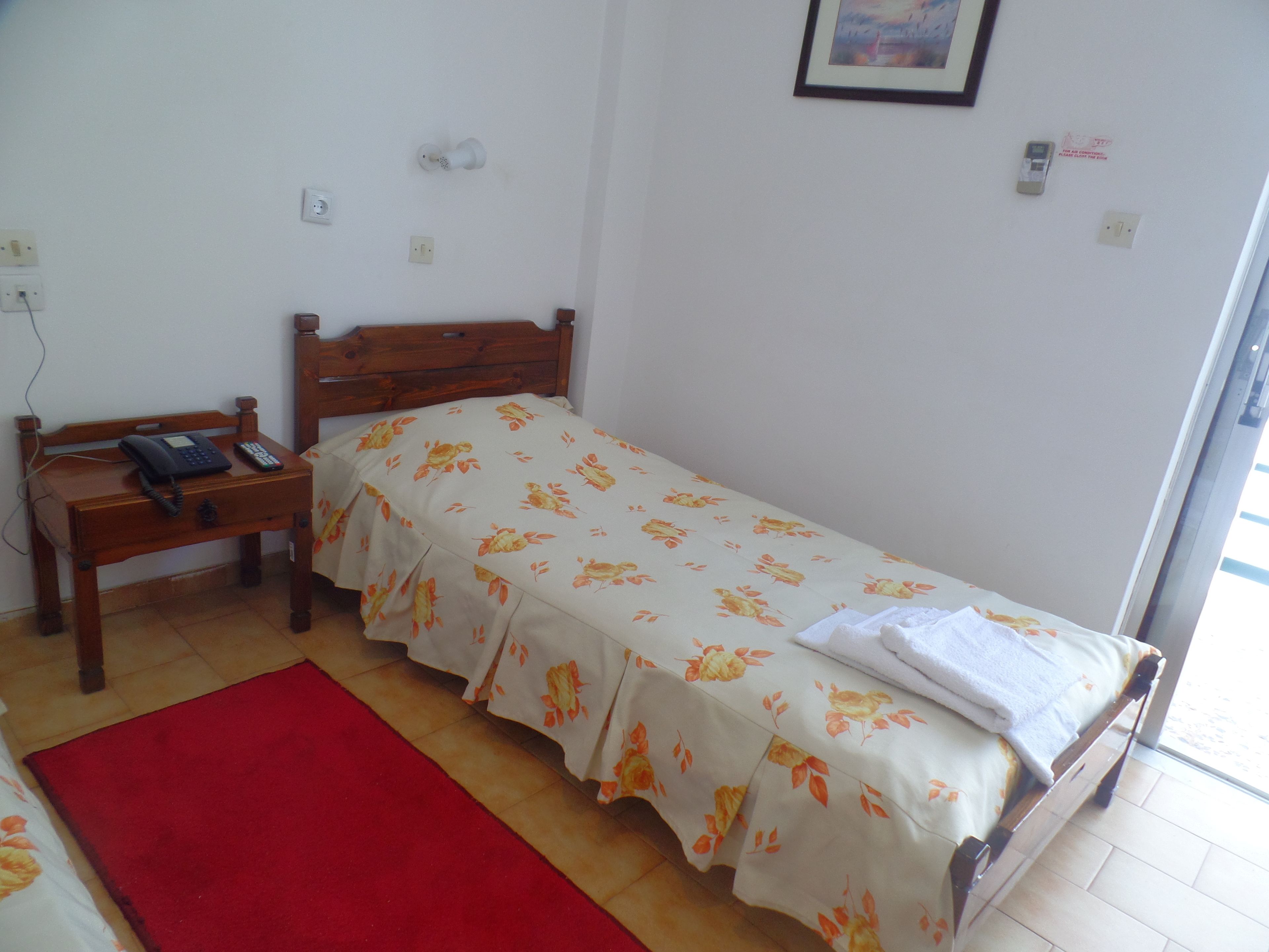 A single-bed room with a wooden headboard, a bedside table with a phone and a lamp, a red rug, and a window with a view of the outside.