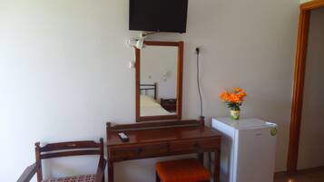 Una habitación con un escritorio de madera, una silla, una mesita con un jarrón de flores, un televisor en la pared y un refrigerador.