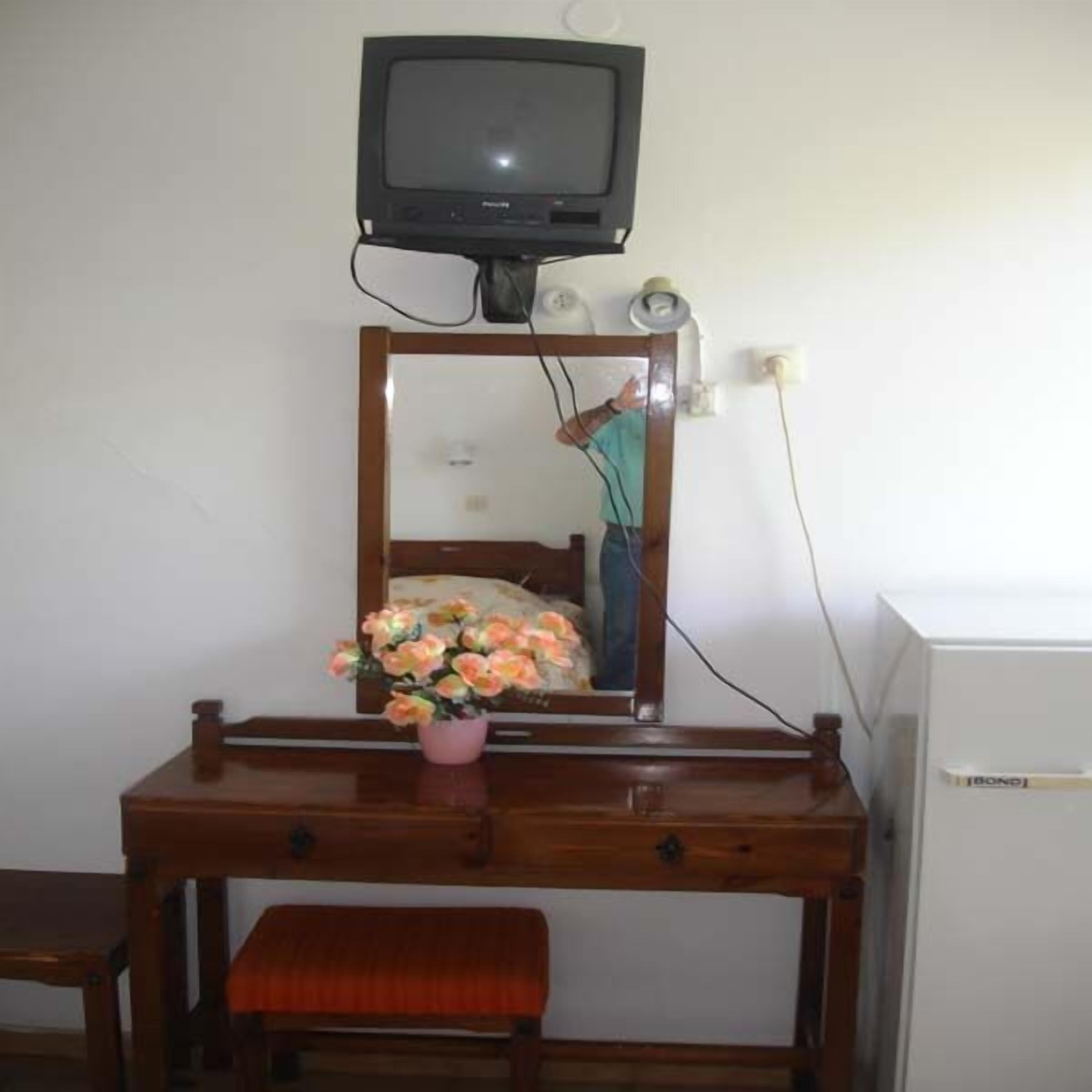 Un bureau en bois avec un téléviseur fixé au-dessus, un miroir et un vase rempli de fleurs.