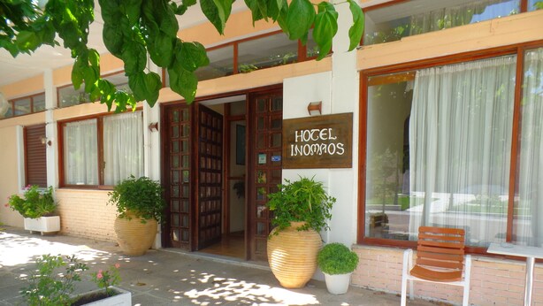 L'ingresso di un hotel con un cartello che riporta la scritta "HOTEL INOMATOS". Sono presenti grandi piante in vaso, un ingresso in pietra e una panca di legno all'esterno.