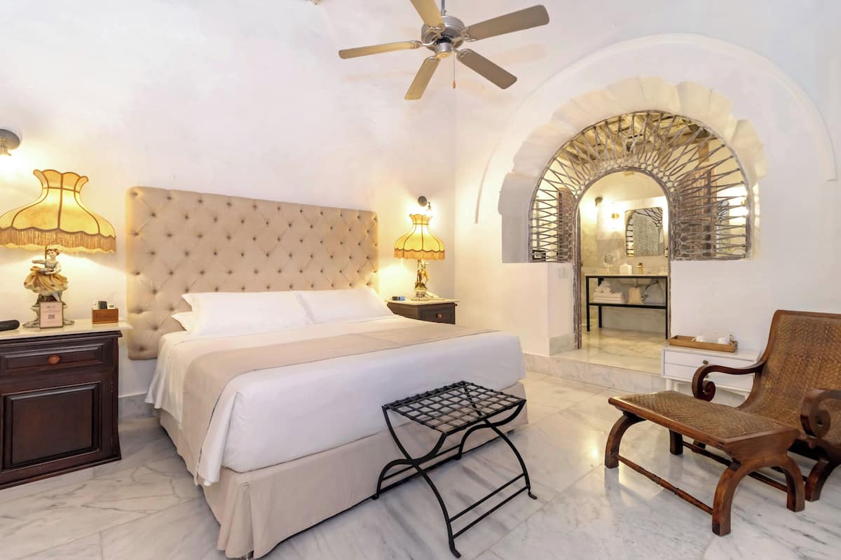 Une chambre à coucher avec une tête de lit capitonnée, un lit recouvert de draps blancs, une table de chevet avec une lampe, une petite table, une chaise et une porte en arc donnant sur une salle de bain.