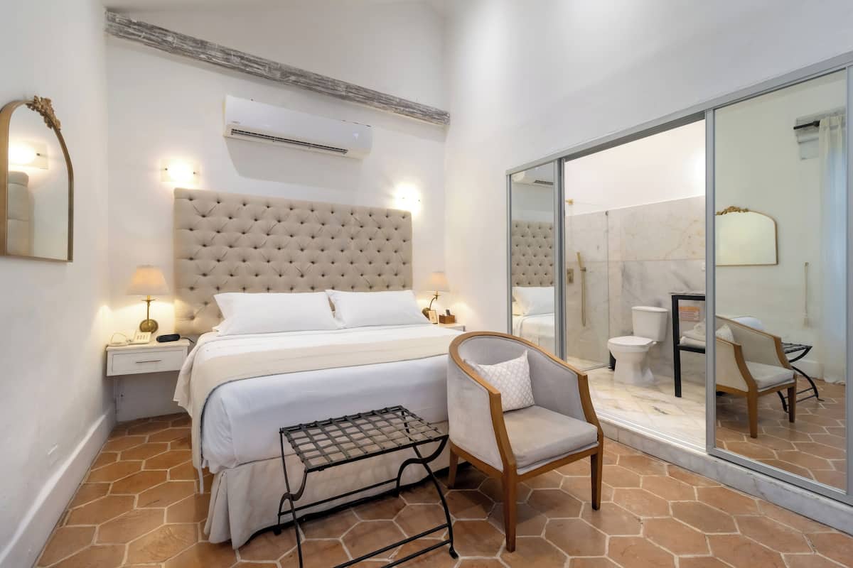 Une chambre à coucher avec une tête de lit capitonnée, un lit avec du linge de lit blanc, une table de chevet avec une lampe, une chaise et une salle de bain fermée par une paroi en verre, équipée de toilettes et d’un lavabo.