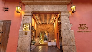 A entrada do Hotel El Marques, com um arco de pedra, duas cadeiras brancas e uma placa para o Bar Restaurante Tito Bravo.