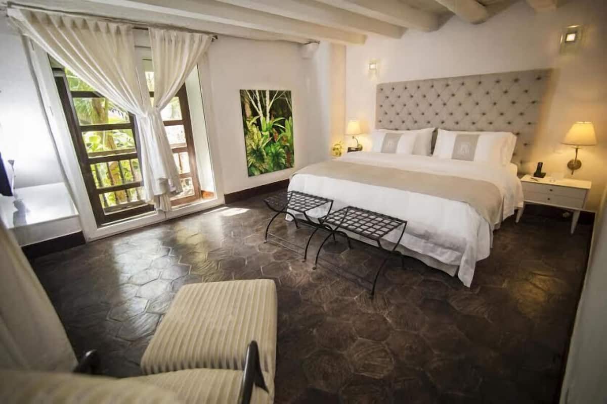 Une chambre à coucher comprenant un lit, une table de chevet, une lampe, une fenêtre avec des rideaux, une chaise et une porte.