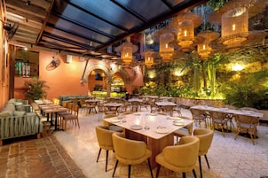 A restaurant with outdoor seating, wooden tables, and chairs, surrounded by a stone wall and greenery.