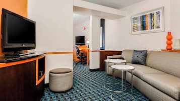 A hotel room with a TV, sofa, two small tables, a patterned carpet, and a framed cityscape picture on the wall.