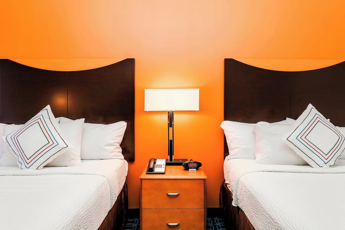 A hotel room with two beds, a nightstand with a lamp, a phone, and a digital clock.