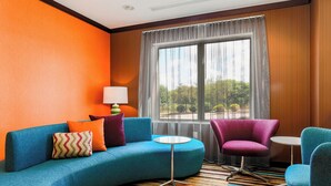 Habitación de hotel moderna con un sofá azul curvo, un sillón púrpura y una pequeña mesa redonda. Un ventanal con cortinas traslúcidas ofrece vistas al verdor exterior.