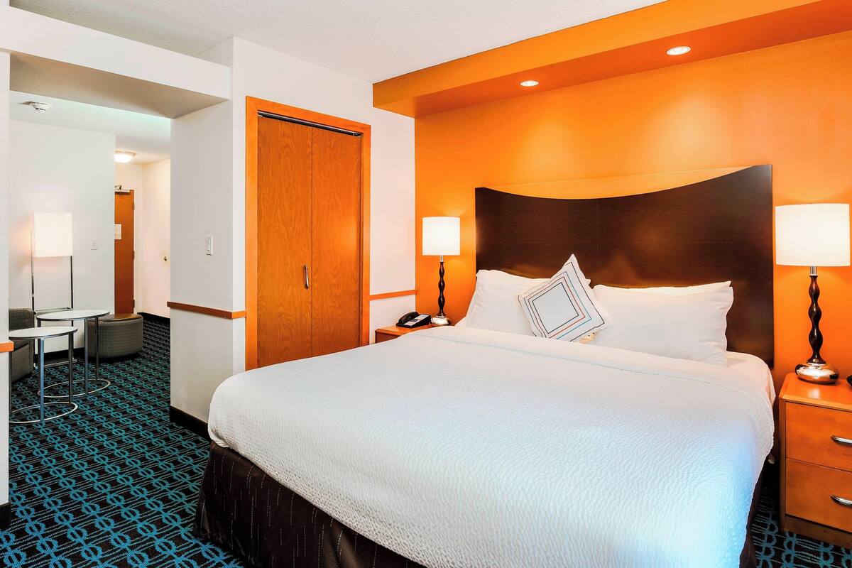 A hotel room with a large bed, a wooden headboard, two bedside tables with lamps, a desk with a chair, and a patterned carpet.