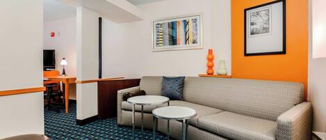 Una habitación de hotel moderna con un sofá gris, dos mesas redondas, un televisor y obras de arte en las paredes.
