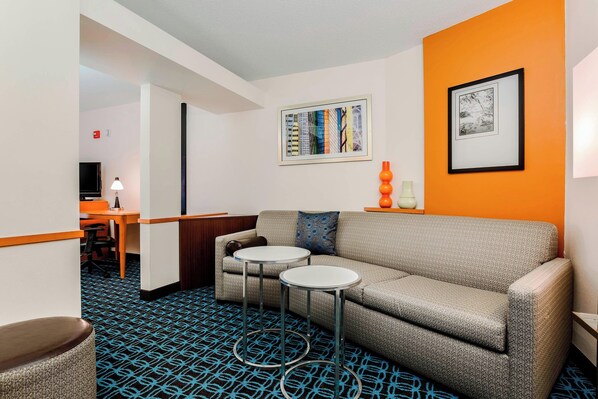 Una habitación de hotel moderna con un sofá gris, dos mesas redondas, un televisor y obras de arte en las paredes.