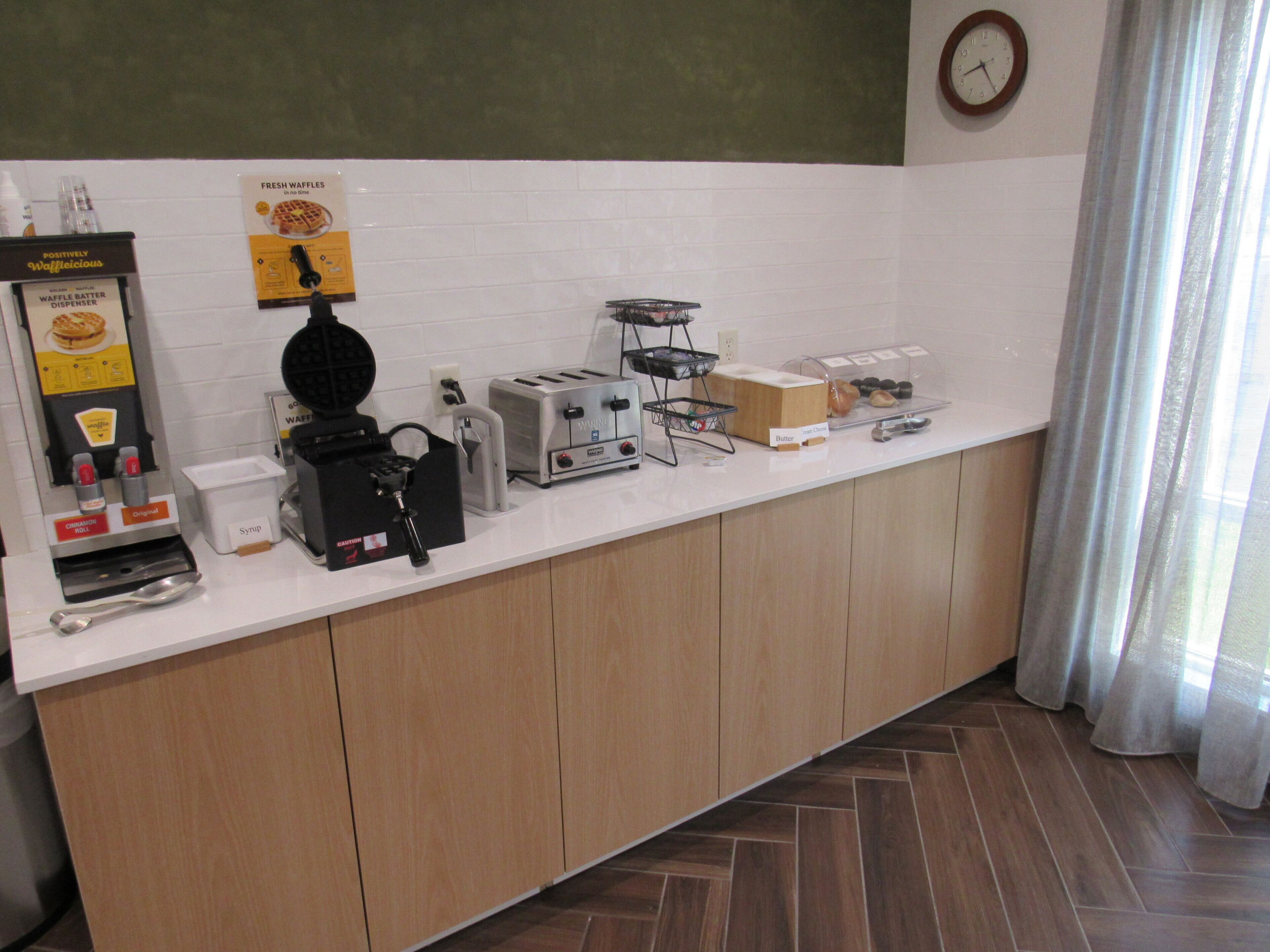 A kitchen counter with waffle batter dispenser, waffle maker, toaster, and butter and jam containers.