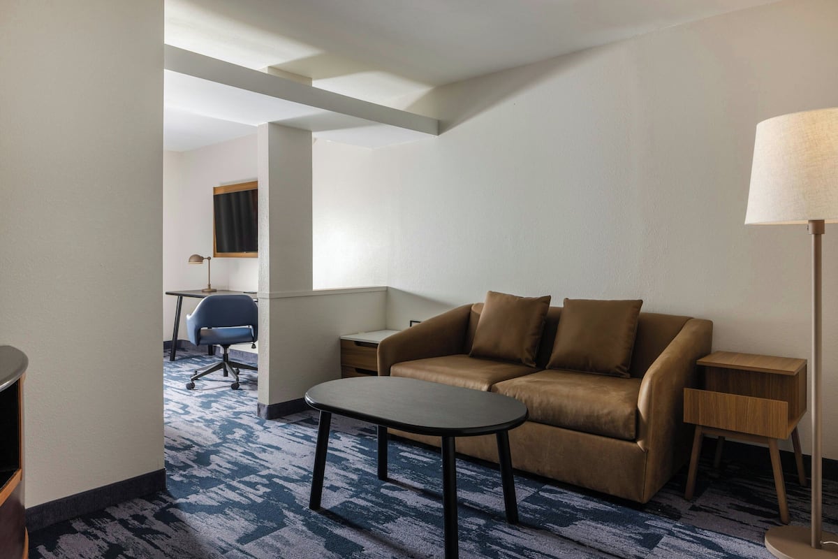 Habitación de hotel con un sofá marrón, una mesita pequeña, una lámpara, un escritorio con una silla y una alfombra estampada.