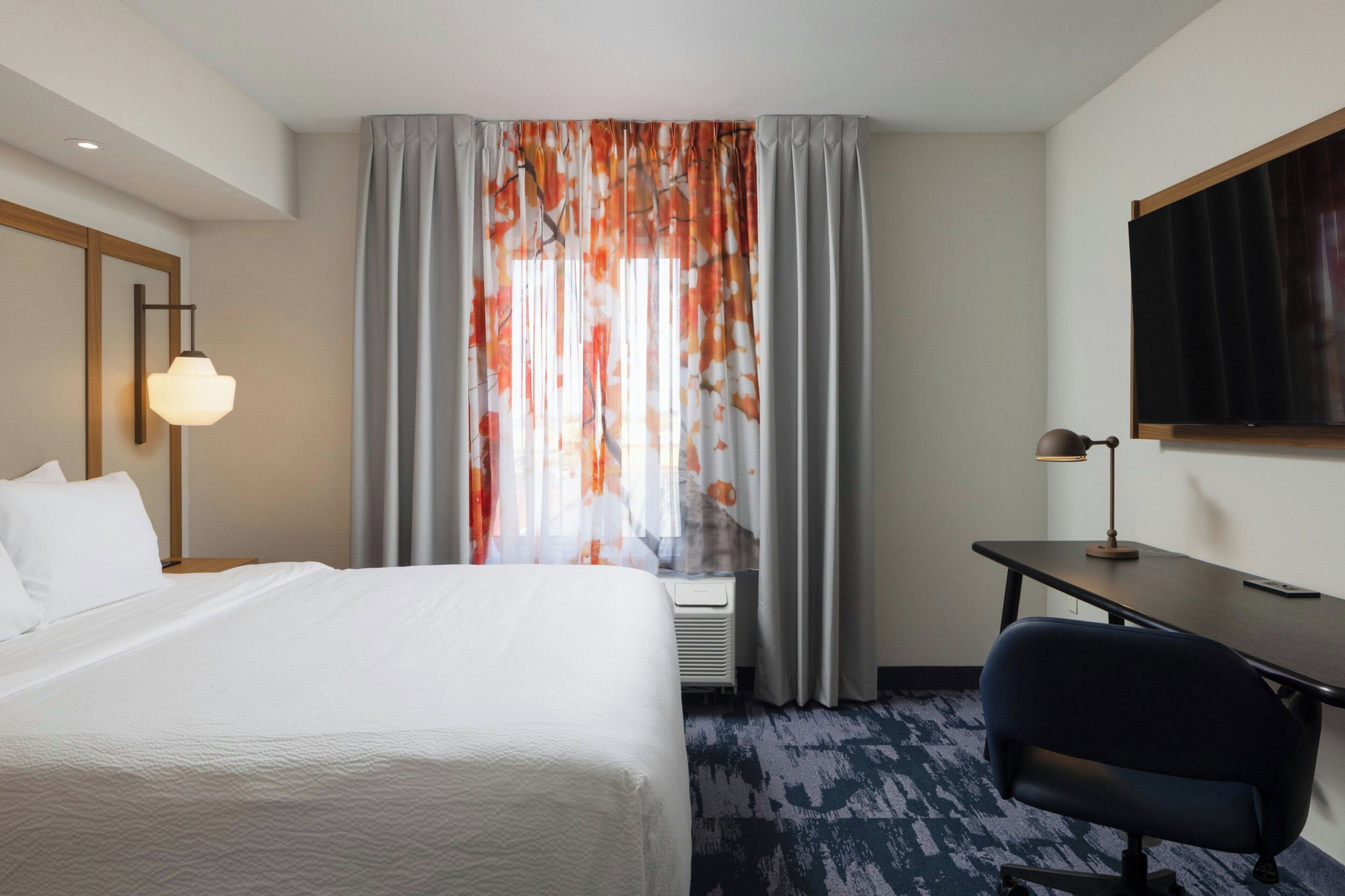 A hotel room with a bed, a desk, a chair, a TV, and a window with curtains.