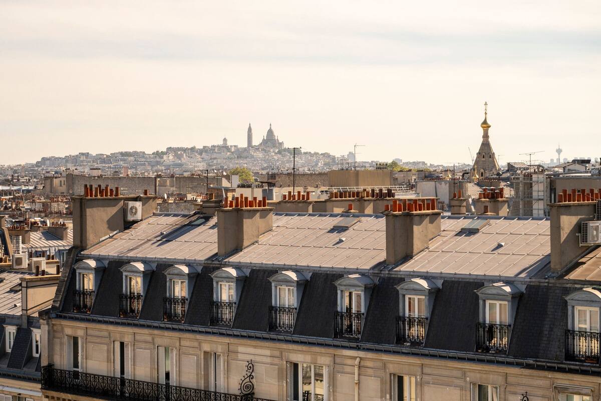 Une vue urbaine avec de nombreux toits, dont celui d’un immeuble doté d’une tour distinctive et, au loin, le clocher d’une église.