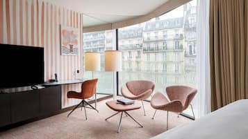 Une chambre d’hôtel dotée d’une grande fenêtre, d’un téléviseur à écran plat, d’un bureau avec une lampe et de deux fauteuils.