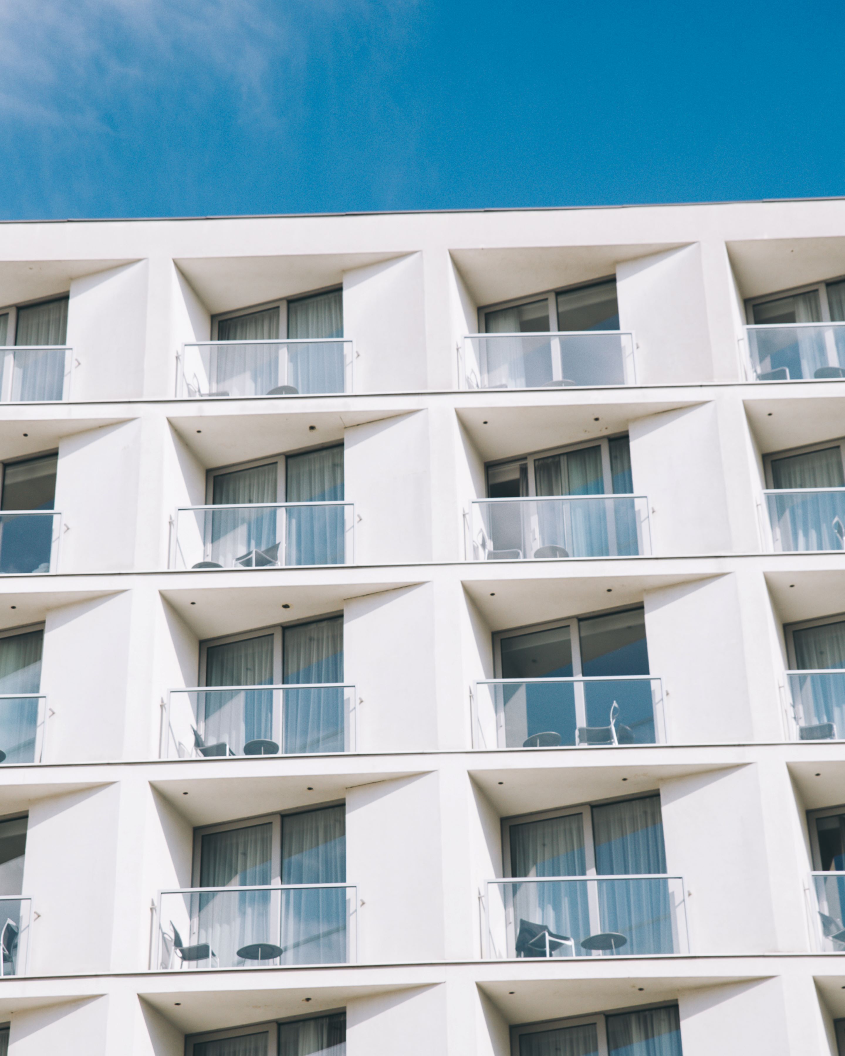 A modern hotel building with multiple balconies, each with a glass railing and a small table and chair.