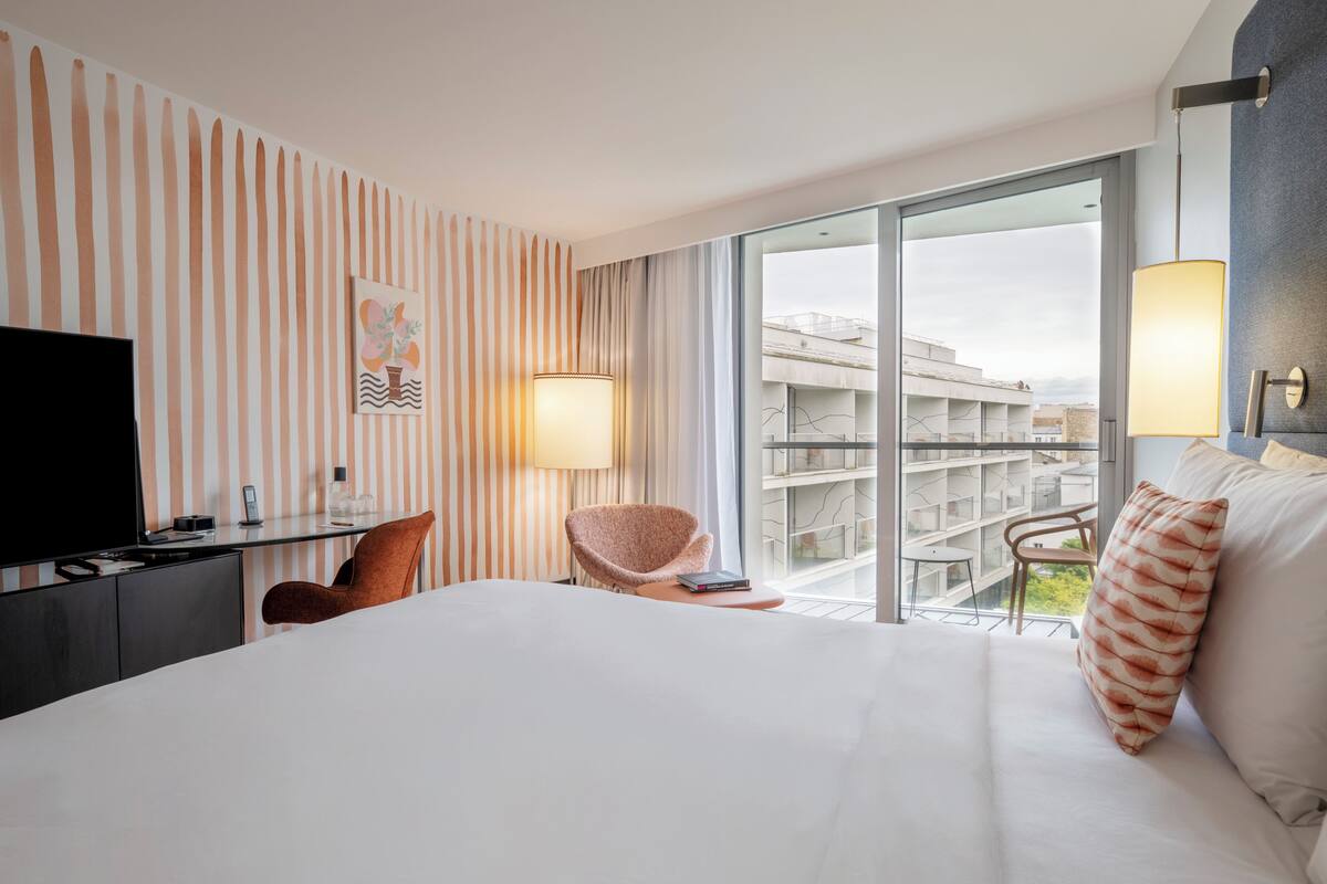 Une chambre d’hôtel équipée d’un lit, d’un bureau, d’une chaise, d’une télévision et d’une grande fenêtre donnant sur des immeubles.
