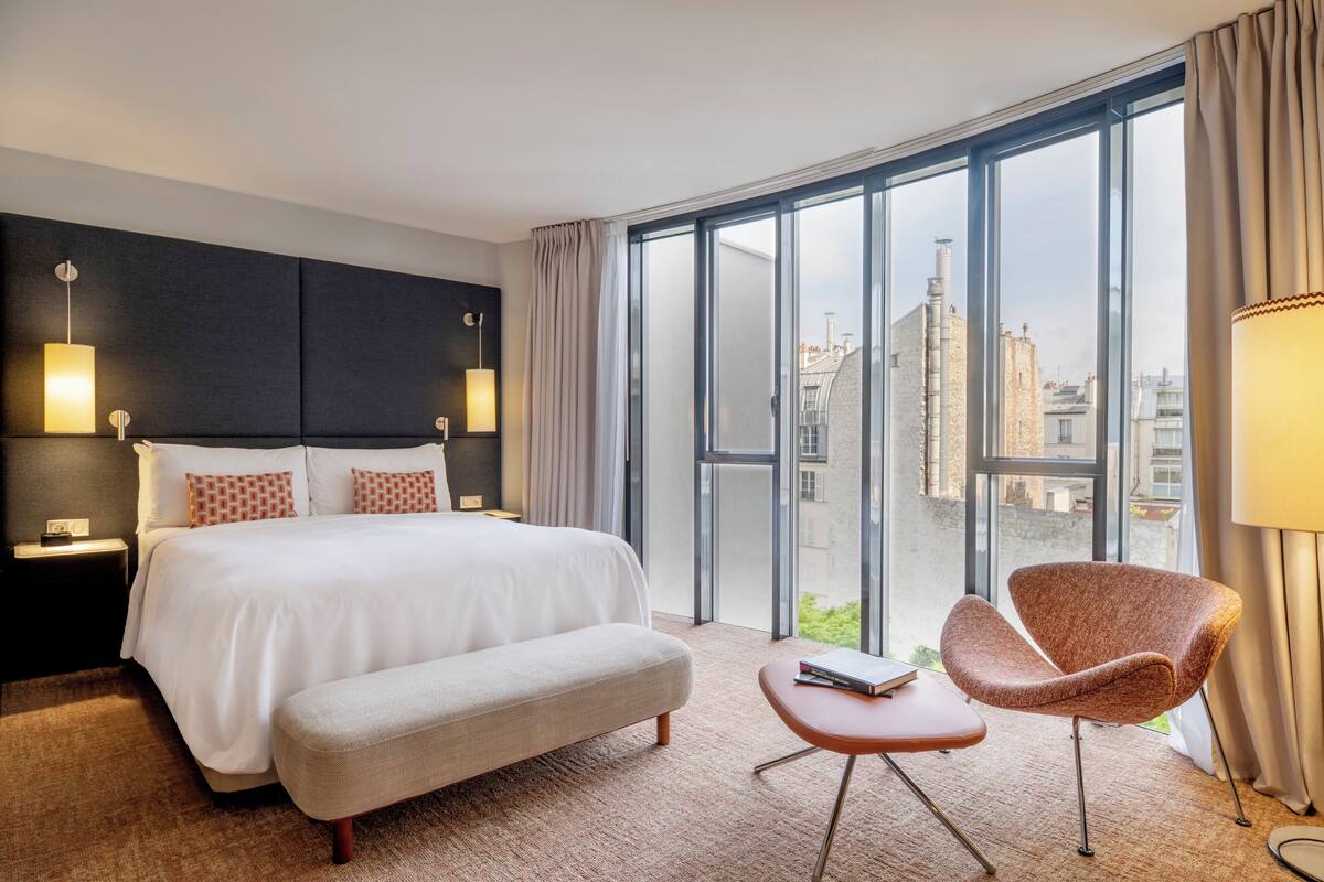 Une chambre d’hôtel avec un grand lit, une banquette, une chaise, une table de chevet et une vue sur la ville grâce à de grandes fenêtres.