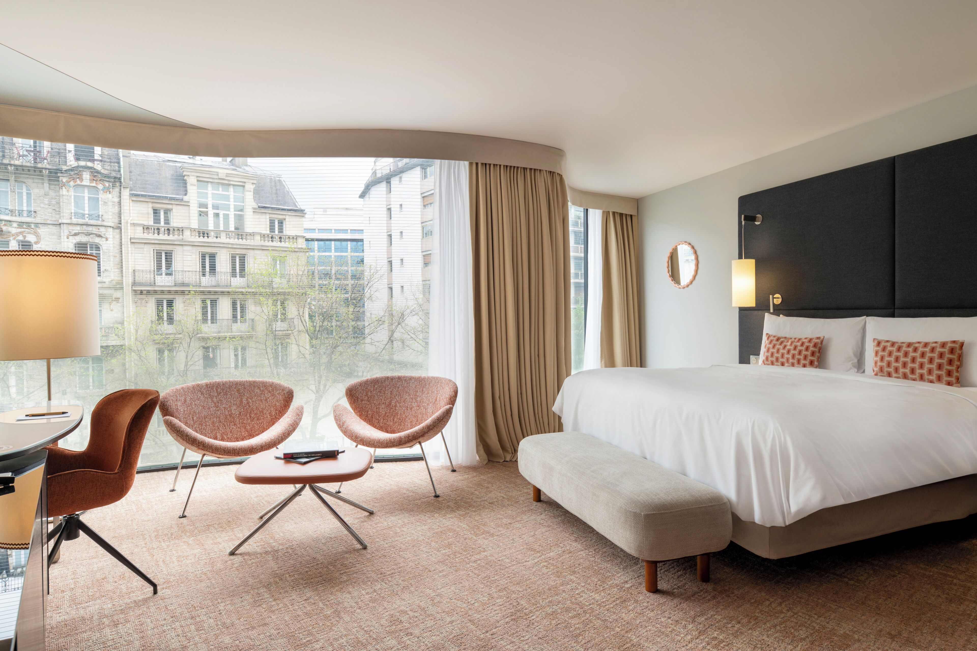 Une chambre d’hôtel comprenant un lit, deux fauteuils, un bureau et une vue sur les bâtiments, grâce à de grandes fenêtres.