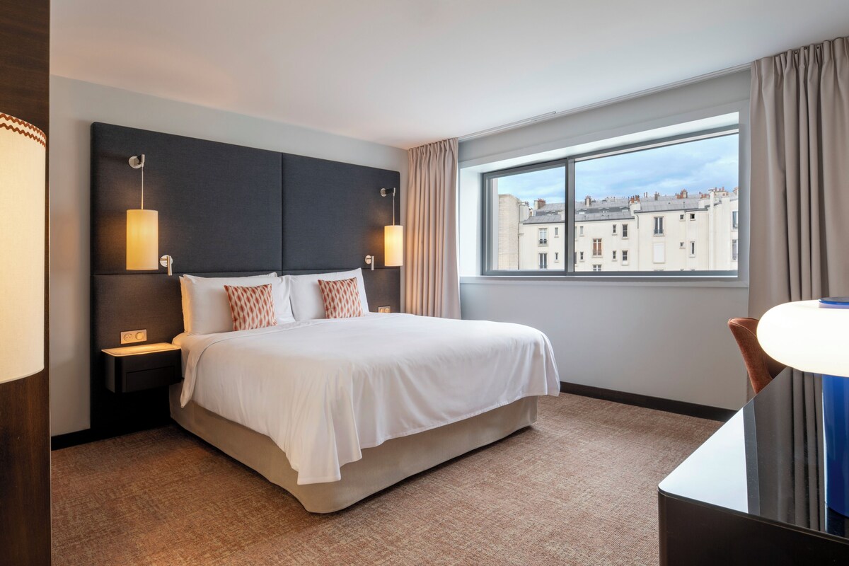 Une chambre d’hôtel avec un grand lit, des lampes de chevet, une vue sur les immeubles par la fenêtre et un bureau avec une lampe.