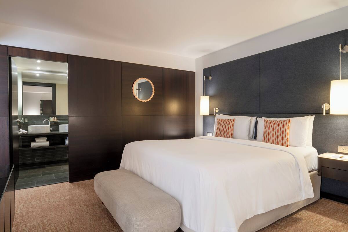 Une chambre d’hôtel moderne avec un grand lit, un miroir rond, une tête de lit sombre et une table de chevet avec une lampe.