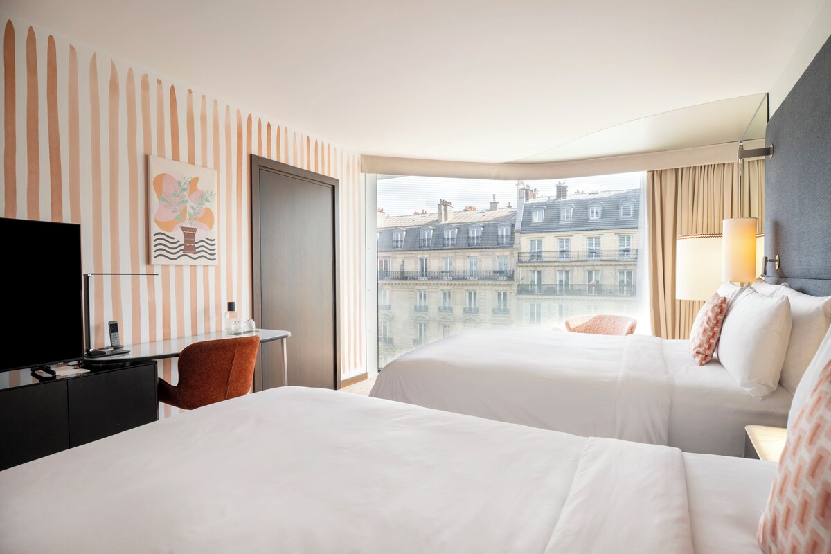 Une chambre d’hôtel avec deux lits, un bureau, une télévision et une vue sur les immeubles.