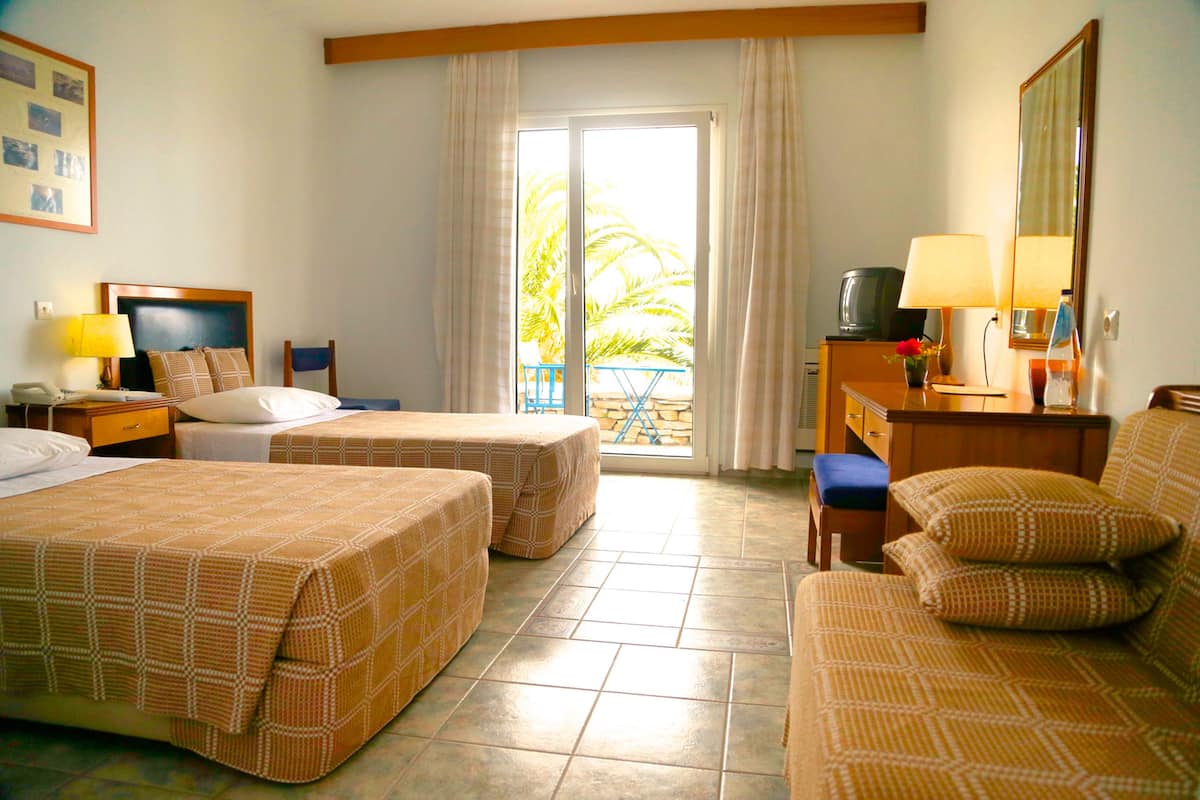 Habitación de hotel con dos camas, un escritorio, una silla, un televisor y un balcón con vistas a palmeras.