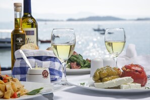 A table with a bottle of wine, two glasses of white wine, a plate of food including stuffed vegetables and cheese, and a view of the sea with boats in the background.