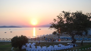 A beachside event with tables and chairs set up for an outdoor gathering, overlooking a calm sea with boats and a sunset in the background. - Xenia (Aristotelis)