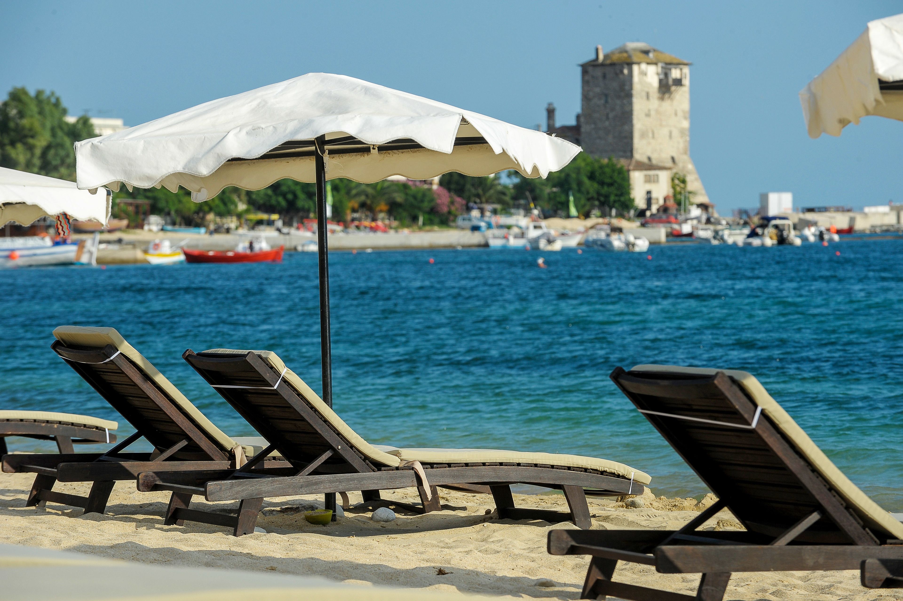A beach with wooden lounge chairs and a white umbrella, a tower in the background, and boats on the water.