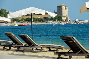 A beach with wooden lounge chairs and a white umbrella, a tower in the background, and boats on the water.