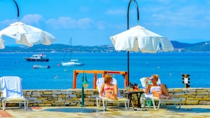 A seaside terrace with two white umbrellas, a stone wall, and a view of boats on the water. - Xenia (Aristotelis)